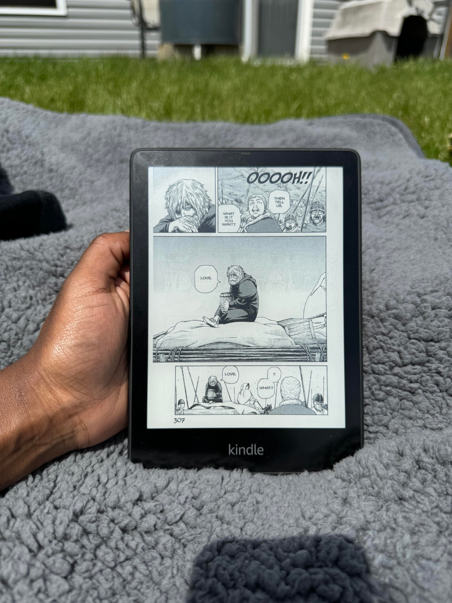 A Kindle resting on a blanket outdoors, showing a full manga page filling the screen.