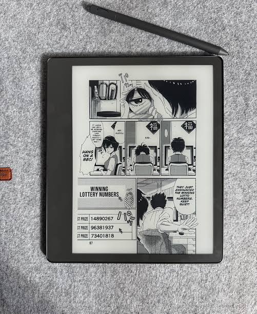 A manga page with dense panels and readable dialogue on an e-ink reader with a stylus beside it.